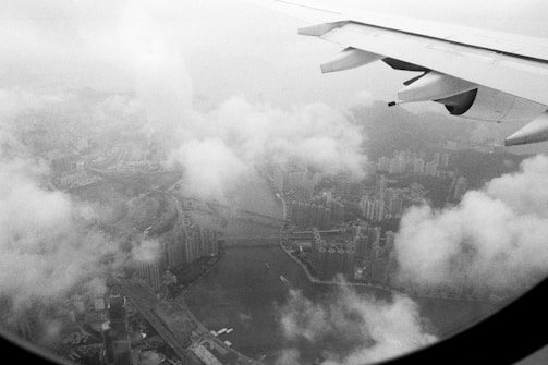 A black and white aerial view of a cityscape covered partly by clouds with an aircraft wing visible in the upper right corner of the image. Skyscrapers and a river or waterway can be seen through the gaps in the clouds.