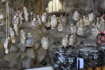 A collection of intricately designed pendants hangs from strings. The pendants feature various carved images and symbols, including faces and the Om symbol. They are displayed over jars filled with black beads, with price tags indicating the cost of the items.