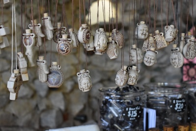 A collection of intricately designed pendants hangs from strings. The pendants feature various carved images and symbols, including faces and the Om symbol. They are displayed over jars filled with black beads, with price tags indicating the cost of the items.