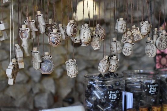 A collection of intricately designed pendants hangs from strings. The pendants feature various carved images and symbols, including faces and the Om symbol. They are displayed over jars filled with black beads, with price tags indicating the cost of the items.