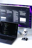 A computer workspace featuring an open laptop displaying code and a larger monitor in the background showing various text-based content. A small figurine resembling a stormtrooper stands on the desk beside the laptop.