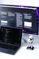 A computer workspace featuring an open laptop displaying code and a larger monitor in the background showing various text-based content. A small figurine resembling a stormtrooper stands on the desk beside the laptop.