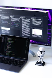 A computer workspace featuring an open laptop displaying code and a larger monitor in the background showing various text-based content. A small figurine resembling a stormtrooper stands on the desk beside the laptop.