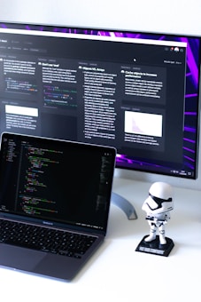 A computer workspace featuring an open laptop displaying code and a larger monitor in the background showing various text-based content. A small figurine resembling a stormtrooper stands on the desk beside the laptop.
