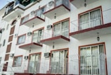 An urban apartment building with a series of symmetrical balconies and large windows. Each balcony has a metal railing and is adorned with an exterior light fixture. The building's facade alternates between sections of white plaster and red brick accents, contributing to a clean, geometric aesthetic. Several air conditioning units are visible on the walls.