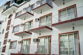 An urban apartment building with a series of symmetrical balconies and large windows. Each balcony has a metal railing and is adorned with an exterior light fixture. The building's facade alternates between sections of white plaster and red brick accents, contributing to a clean, geometric aesthetic. Several air conditioning units are visible on the walls.