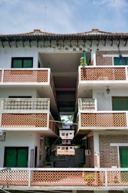 A multi-story residential building with a tiled roof. The structure features multiple balconies with decorative lattice railings. The facade is a mix of white and exposed brick sections, with vibrant green window shutters. The open central passage offers a view of additional similar buildings in the background.