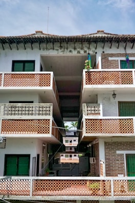 A multi-story residential building with a tiled roof. The structure features multiple balconies with decorative lattice railings. The facade is a mix of white and exposed brick sections, with vibrant green window shutters. The open central passage offers a view of additional similar buildings in the background.