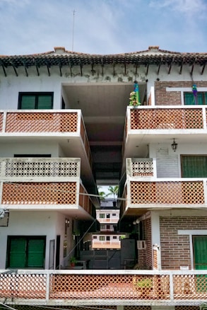 A multi-story residential building with a tiled roof. The structure features multiple balconies with decorative lattice railings. The facade is a mix of white and exposed brick sections, with vibrant green window shutters. The open central passage offers a view of additional similar buildings in the background.