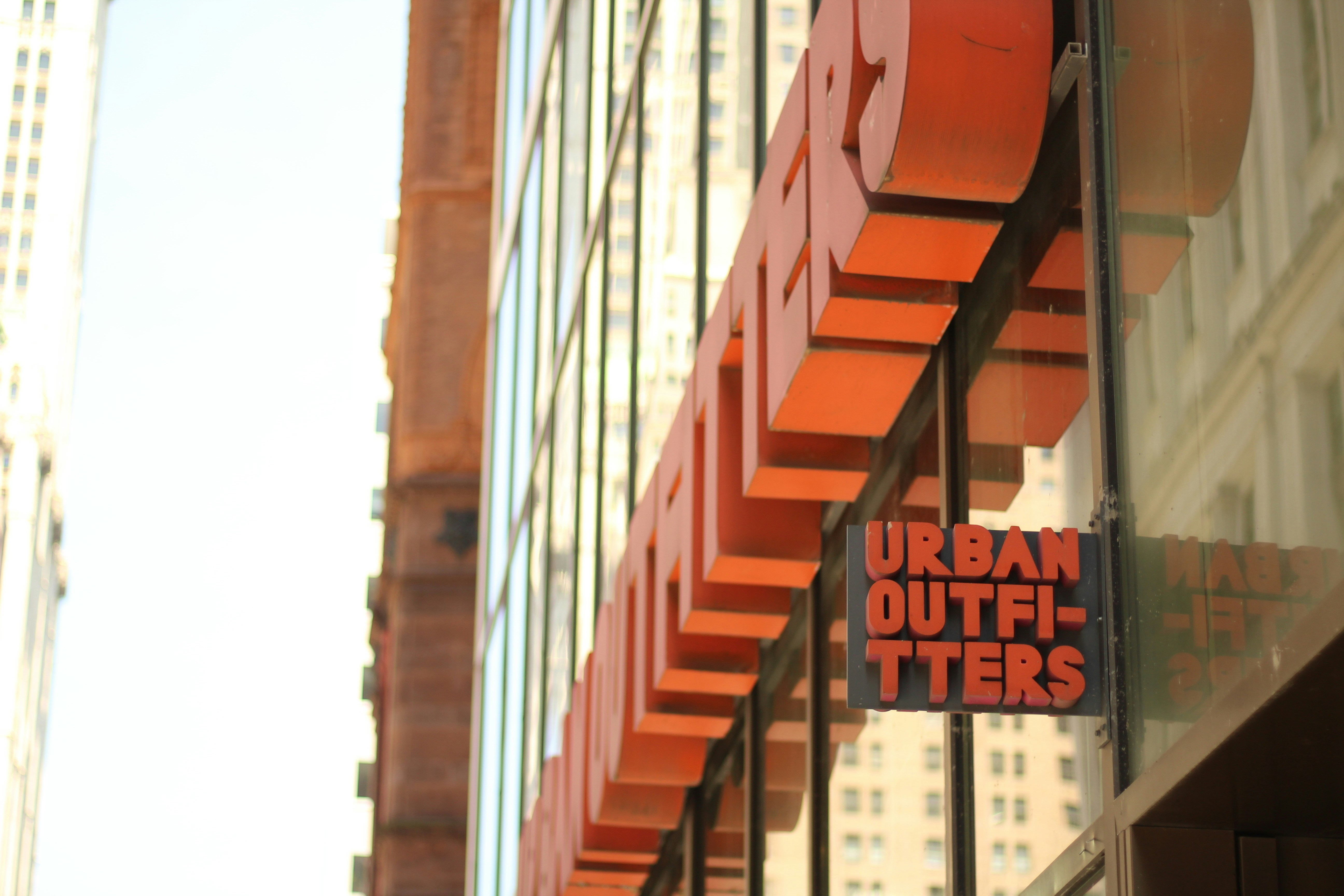 A large glass storefront features a prominent Urban Outfitters sign in bold, orange letters. The building reflects nearby urban architecture with tall windows and minimalistic design.