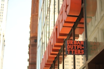 A large glass storefront features a prominent Urban Outfitters sign in bold, orange letters. The building reflects nearby urban architecture with tall windows and minimalistic design.