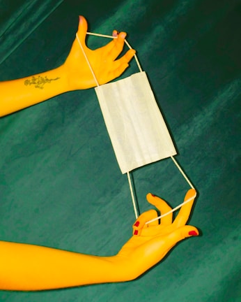 Two hands with painted nails are holding a white face mask by its ear loops against a dark green background. One hand features a floral tattoo.