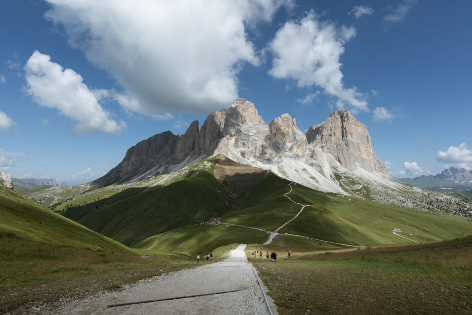 The image depicts a picturesque mountain range with three prominent peaks, each varying in height and color - one white, one gray, and one brown. The mountains are nestled amidst lush green valleys, creating a striking contrast between the natural beauty of the landscape and the man-made structures below. A winding road meanders through the valley, leading up to the majestic peaks. Several people can be seen walking along this path, adding a sense of scale and activity to the scene.

The sky above is a clear blue with fluffy white clouds scattered across it, providing a serene backdrop for the mountain range below. The image is taken from a low angle, looking up at the mountains, which enhances their grandeur and prominence in the landscape.