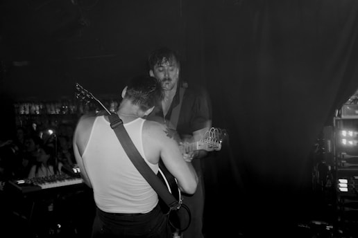Two musicians are on stage, deeply engaged in a live performance. One of them plays an electric guitar while the other, wearing a sleeveless shirt, leans in. The scene captures the intimacy and energy of a live music show, with dim lighting and audience presence partially visible in the background.