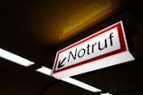An emergency sign with the word 'Notruf' in bold black letters on a white background, outlined with a red border. The sign features a black arrow pointing left and is mounted on a ceiling with perforated panels. Fluorescent lights are visible, casting an industrial ambiance.