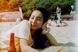 A relaxed person lies on the sand at the beach with a content expression, resting their head on their hand. In the foreground, there is a bottle on the sand. In the background, a few people are seen walking, while a white building and some trees are visible.