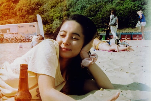 A relaxed person lies on the sand at the beach with a content expression, resting their head on their hand. In the foreground, there is a bottle on the sand. In the background, a few people are seen walking, while a white building and some trees are visible.