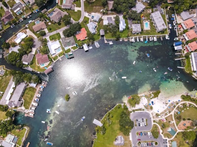 An aerial view of a waterfront residential area with houses lined along a canal. Several small boats are docked, and others are moving through the water. A public beach area with people and parked cars is visible on the right, surrounded by greenery.