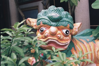 A colorful, decorative statue resembling a mythical creature, partially obscured by green foliage. The creature has a vibrant mix of colors with distinct features such as large, expressive eyes and a patterned body.