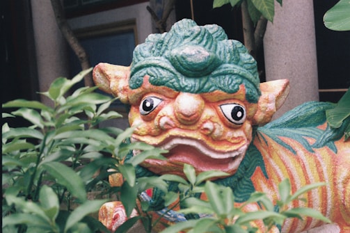 A colorful, decorative statue resembling a mythical creature, partially obscured by green foliage. The creature has a vibrant mix of colors with distinct features such as large, expressive eyes and a patterned body.
