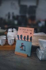 An illustrated card is displayed on a wooden stand featuring a stylized person riding a scooter against a backdrop of trees and an orange sky. The text 'AS FAST AS THE WIND' is printed above the illustration. The image is set in a cozy, dimly lit environment with teacups on a wooden tray and a box of tissues visible nearby.
