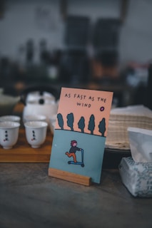 An illustrated card is displayed on a wooden stand featuring a stylized person riding a scooter against a backdrop of trees and an orange sky. The text 'AS FAST AS THE WIND' is printed above the illustration. The image is set in a cozy, dimly lit environment with teacups on a wooden tray and a box of tissues visible nearby.