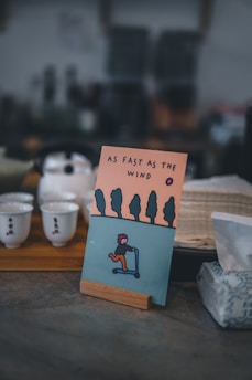 An illustrated card is displayed on a wooden stand featuring a stylized person riding a scooter against a backdrop of trees and an orange sky. The text 'AS FAST AS THE WIND' is printed above the illustration. The image is set in a cozy, dimly lit environment with teacups on a wooden tray and a box of tissues visible nearby.