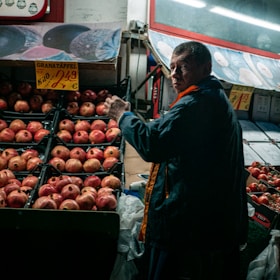A person is standing in front of a display of pomegranates in a market setting. There are signs with prices above the fruits, and the lighting is somewhat dim, suggesting an indoor environment or evening time.
