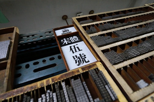 Wooden drawers filled with metal type pieces used for printing are organized in rows. There is a signboard with Chinese characters in the center and metal shelving visible.