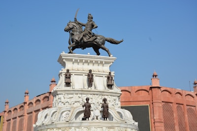 A statue of a mounted warrior wielding a sword dominates the scene. The monument is ornate, with additional sculptures of people around its base. Behind the statue is a building made of red stone with arched designs.