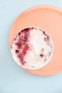 A close-up view of a cup containing a creamy dessert with swirls of red and white, indicating a mix of ice cream or yogurt with fruit or berry sauce. The cup is set on a peach-colored plate positioned against a light blue background, creating a soft, pastel aesthetic.