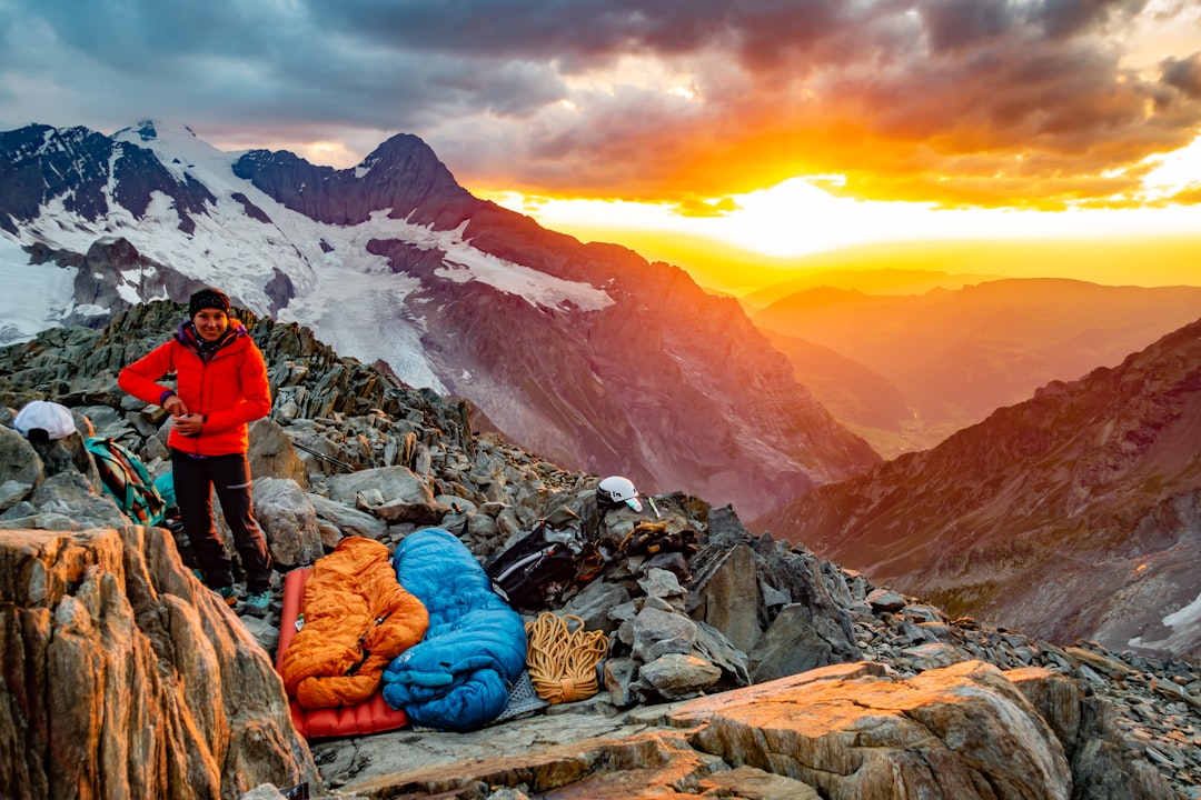 person in red jacket and blue denim jeans sitting on rock near snow covered mountain during