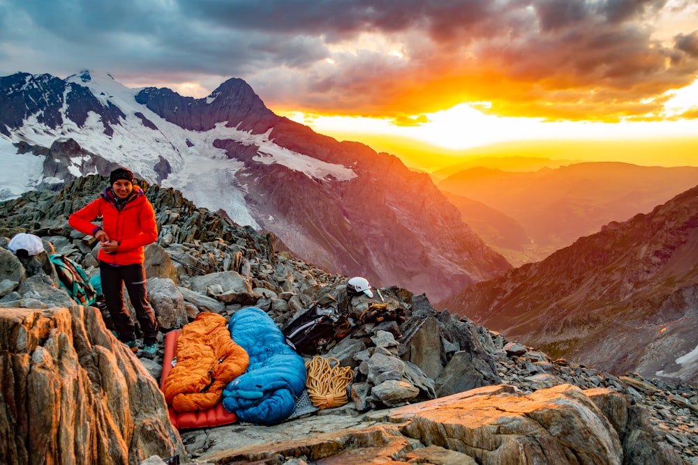 person in red jacket and blue denim jeans sitting on rock near snow covered mountain during