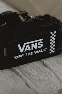 A black camera displaying the Vans branding with the phrase 'Off The Wall' and a distinctive checkered pattern, placed on a textured fabric surface.