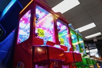 A row of vibrant claw machines line an indoor arcade, each adorned with brightly lit displays and priced at 50 cents. The foreground machine is predominantly red, with others in different colors, all under fluorescent ceiling lights.