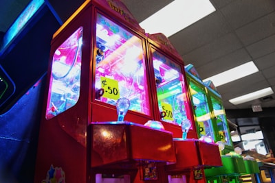 A row of vibrant claw machines line an indoor arcade, each adorned with brightly lit displays and priced at 50 cents. The foreground machine is predominantly red, with others in different colors, all under fluorescent ceiling lights.