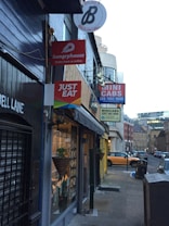 A narrow urban street with several restaurant and service signs hanging on the buildings. Signs include 'hungryhouse', 'JUST EAT', and 'MINI CABS'. The storefronts have dark exteriors, and there is a view of cars parked on the side of the street. A decorative plant hangs near one of the windows, and the street is lined with cobblestones.