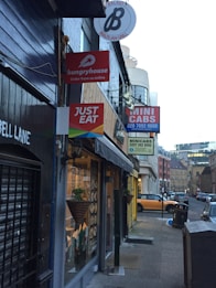 A narrow urban street with several restaurant and service signs hanging on the buildings. Signs include 'hungryhouse', 'JUST EAT', and 'MINI CABS'. The storefronts have dark exteriors, and there is a view of cars parked on the side of the street. A decorative plant hangs near one of the windows, and the street is lined with cobblestones.