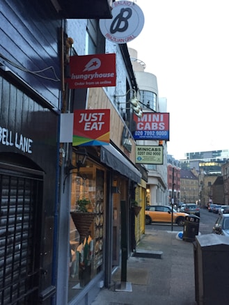 A narrow urban street with several restaurant and service signs hanging on the buildings. Signs include 'hungryhouse', 'JUST EAT', and 'MINI CABS'. The storefronts have dark exteriors, and there is a view of cars parked on the side of the street. A decorative plant hangs near one of the windows, and the street is lined with cobblestones.