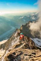 Wide-angle view of a climber reaching a summit with expansive sky and mountains behind.