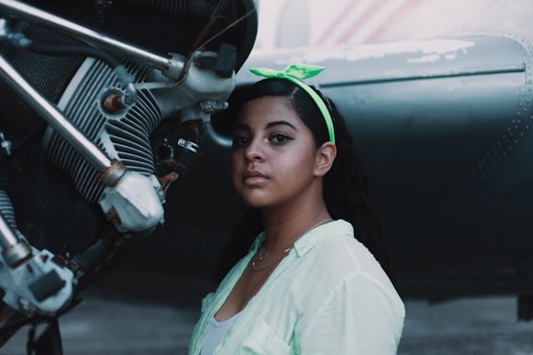 A person stands next to a mechanical engine with intricate components. The individual wears a green headband and a light-colored shirt, looking directly at the camera. The setting appears industrial, with metallic elements and a soft lighting ambiance.