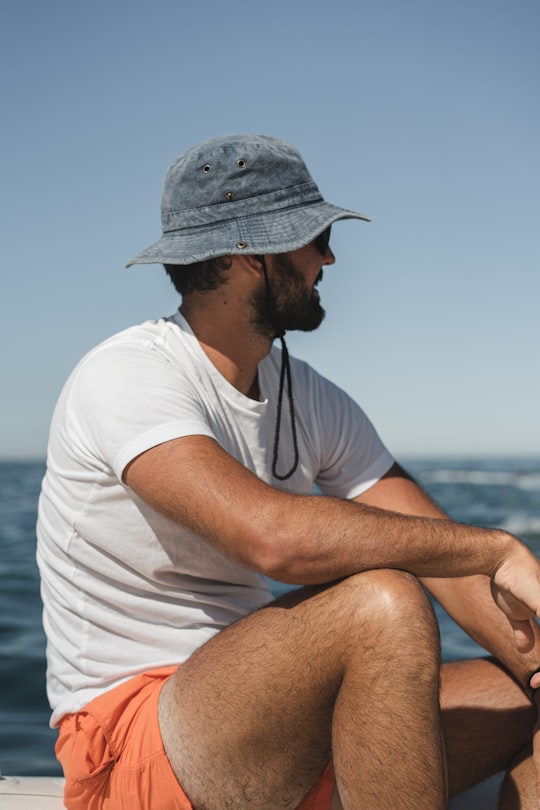 man in white t-shirt and gray fedora hat sitting on brown rock during daytime in La Baule France