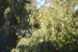 A dream catcher hangs against a blurred background of lush green foliage. The dream catcher features a circular frame with a web-like pattern in the center and three feathers dangling from its bottom.