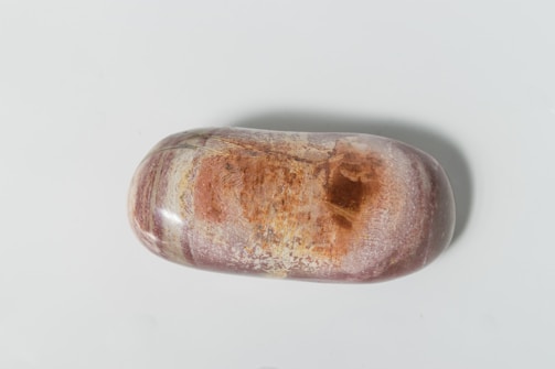 A polished, oval-shaped stone with a smooth surface. The stone features a mix of reddish and brownish hues with streaks of cream and white. It has a glossy finish, highlighting the natural patterns on its surface.