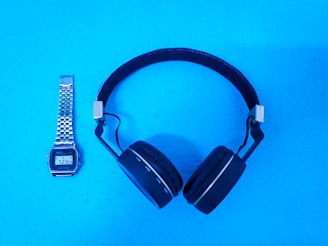 A pair of over-ear headphones and a digital wristwatch are placed on a bright blue surface. The headphones are black with a metallic adjustment piece, and the wristwatch has a silver metallic strap and a digital display showing the time as 7:23.