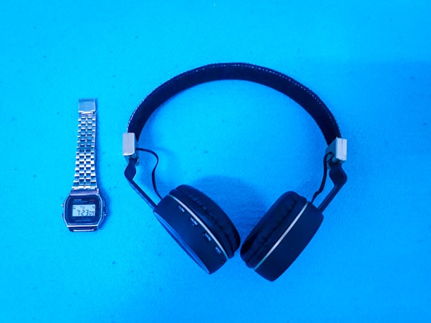 A pair of over-ear headphones and a digital wristwatch are placed on a bright blue surface. The headphones are black with a metallic adjustment piece, and the wristwatch has a silver metallic strap and a digital display showing the time as 7:23.