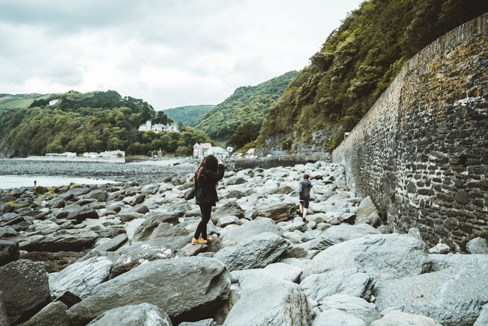 uomo in giacca nera e pantaloni gialli in piedi sul fiume roccioso durante il giorno