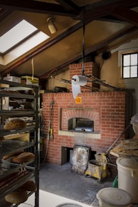 A brick oven setup in a bakery with a rustic interior. Baked goods are placed on metal racks to the left, and a roll of paper towel hangs from the ceiling. The space is lit through a skylight and a small window, casting light on the oven area, which has various baking tools and supplies around it.