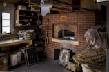 A rustic kitchen with a brick pizza oven is surrounded by various kitchen items. Stacks of baking trays and bowls are on shelving, and a large roll of paper towels hangs from the ceiling. The counter is cluttered with baking essentials and reflective metal containers. The overall setup conveys a functional, slightly cluttered baking workspace.
