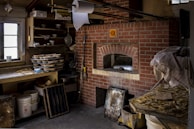A rustic kitchen with a brick pizza oven is surrounded by various kitchen items. Stacks of baking trays and bowls are on shelving, and a large roll of paper towels hangs from the ceiling. The counter is cluttered with baking essentials and reflective metal containers. The overall setup conveys a functional, slightly cluttered baking workspace.